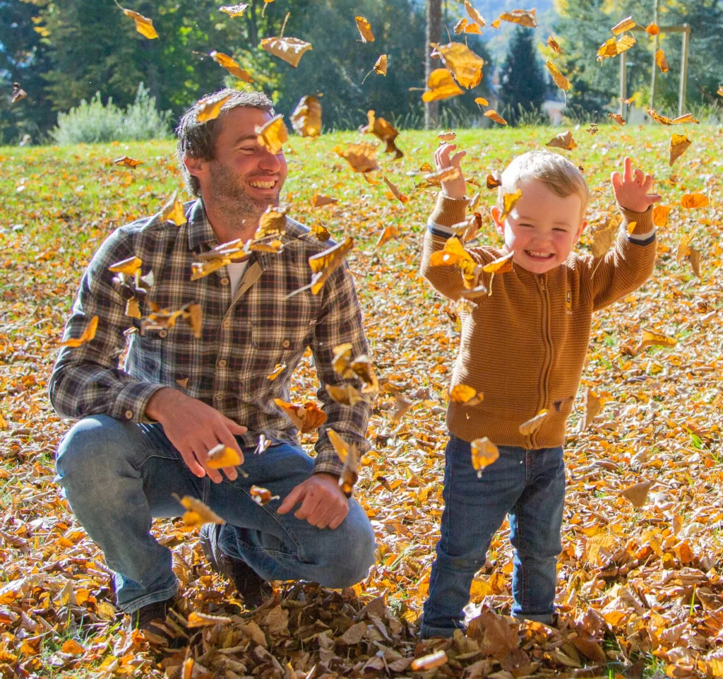Photo d’un père et de son fils jouant avec des feuilles en automne, moment de complicité et de bonheur capturé en lumière naturelle dans les Hautes-Pyrénées
