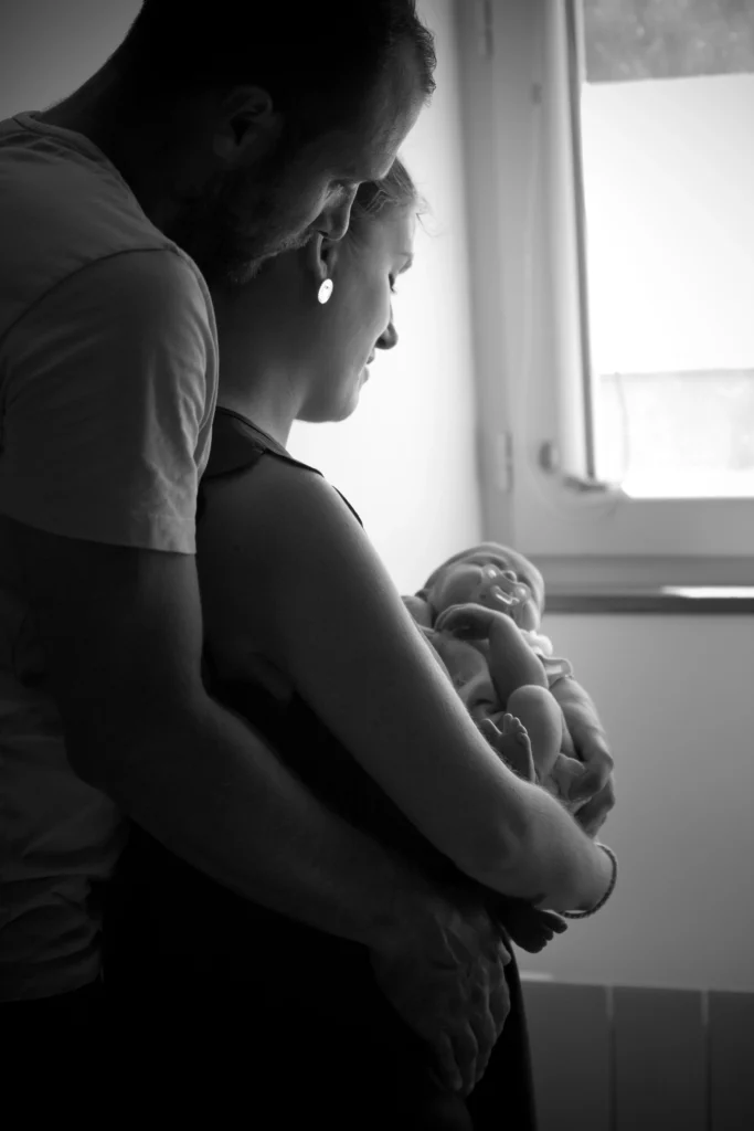 Photo de naissance en noir et blanc d’un couple tenant leur bébé dans les bras près d’une fenêtre à Azereix, réalisée par LS Productions photographe naissance